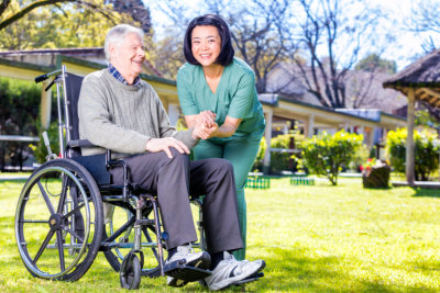 Asian nurse happy with caucasian elder patient on wheelchair, outdoor view.